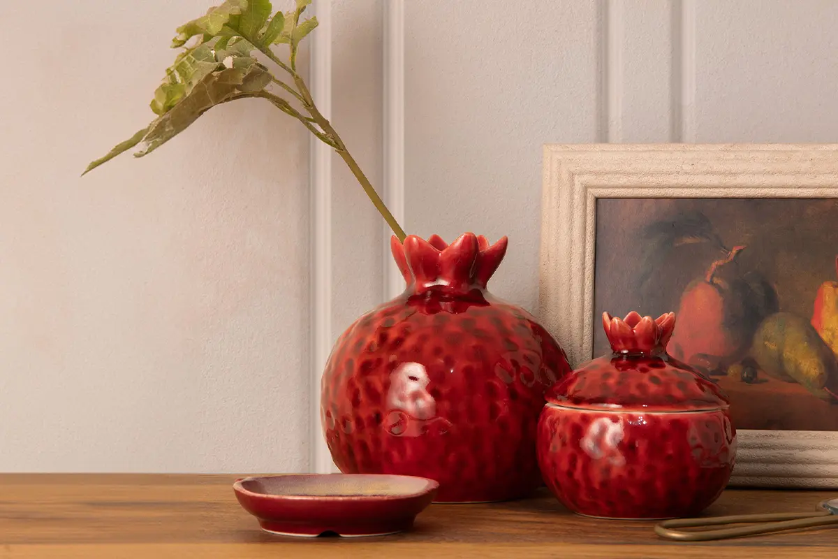 Mesa de madera con florero y contenedor con tapa de cerámica roja en forma de granada, junto a un cuadro decorativo con marco madera