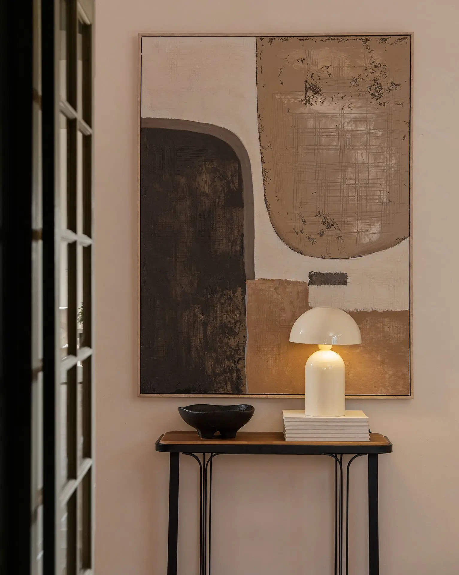 Mesa consola de madera y metal con una lámpara de mesa moderna encendida sobre libros, un bowl decorativo negro y un cuadro abstracto grande en tonos tierra y neutros de fondo.